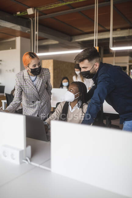 Group of diverse coworkers in protective masks gathering on laptop and discussing project while working together in contemporary workspace — Stock Photo