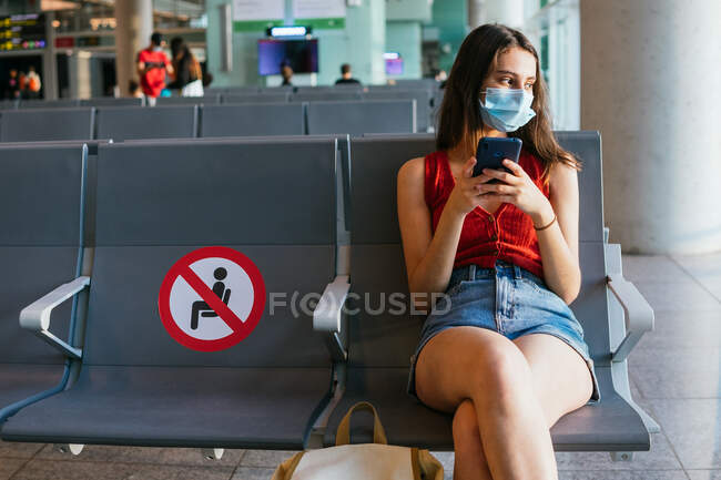 Turista con máscara protectora sentada en la sala de salida del aeropuerto y esperando el vuelo durante la epidemia de coronavirus mientras usa el teléfono inteligente - foto de stock