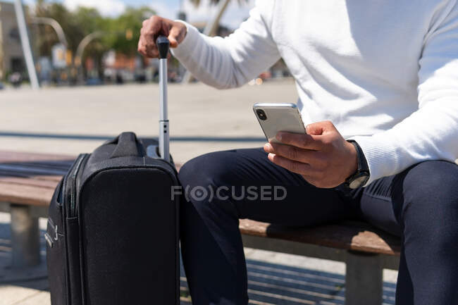 Crop male in smart casual clothes browsing mobile phone and sitting on sunny street with suitcase — Stock Photo