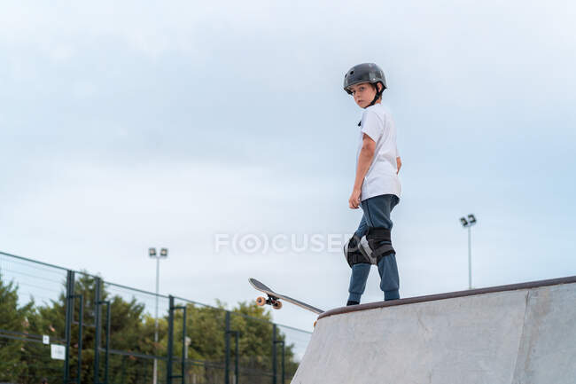 Pattinatore adolescente in attrezzatura protettiva guida skateboard durante il fine settimana in skate park e guardando altrove — Foto stock