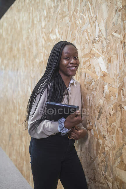 Mujer empresaria afroamericana sonriente de pie con tableta cerca de la pared en el espacio de coworking mientras mira la cámara - foto de stock