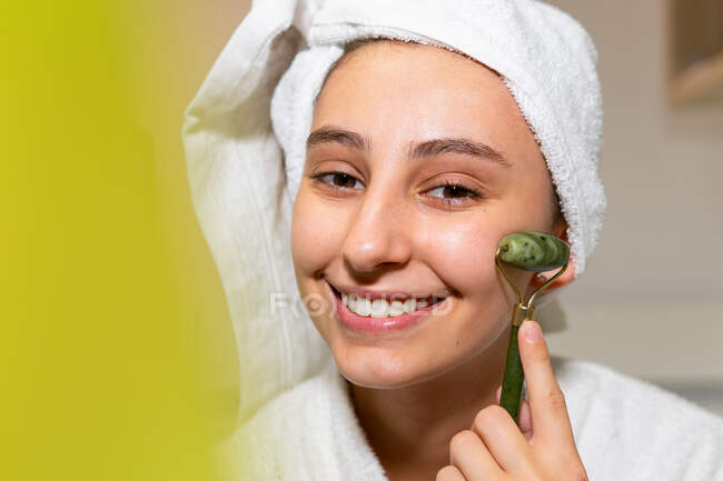 Feliz joven hembra con toalla en la cabeza sonriendo y masajeando la cara con rodillo de jade durante la rutina de cuidado de la piel en casa - foto de stock