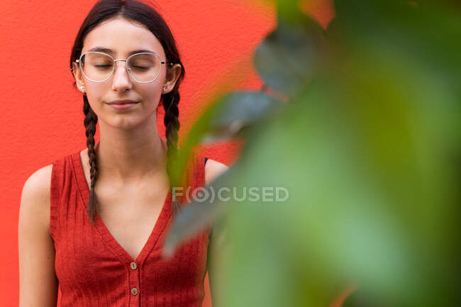 Молода безтурботна жінка в стильних окулярах, що стоять на червоному тлі на вулиці з зеленими рослинами — стокове фото