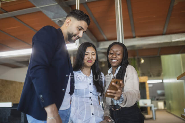 Low angle of group of happy multiethnic colleagues standing in contemporary coworking space and watching video on mobile phone during break — Stock Photo