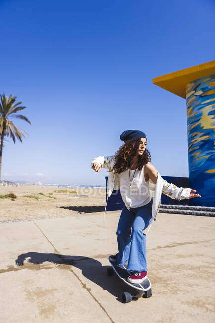Повне тіло активної жінки в повсякденному одязі катається на скейтборді по дорозі вздовж піщаного пляжу і високих долонь під час тренувань, дивлячись далеко — стокове фото