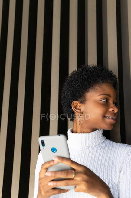Glückliche junge schwarze Frau im Pullover surft auf dem Smartphone, während sie in der Nähe einer gestreiften Hauswand in der Stadt steht — Stockfoto