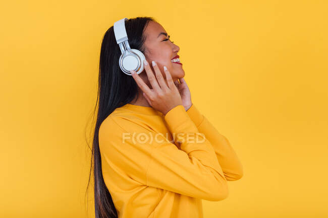 Vista lateral de una mujer asiática positiva disfrutando de canciones y escuchando música en auriculares sobre fondo amarillo en el estudio - foto de stock