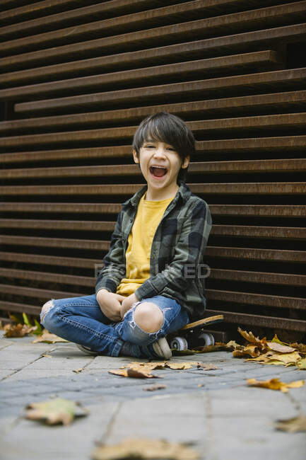 Full body of smiling boy in casual clothes looking at camera while sitting on skateboard at wall on street with fallen leaves — Stock Photo