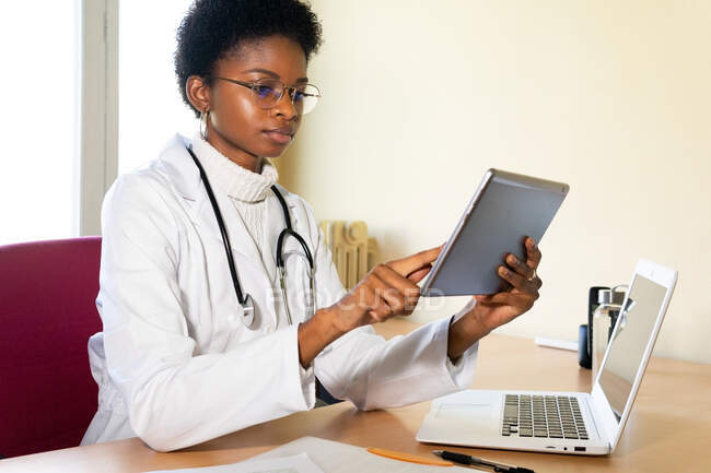 Jovem médica negra em casaco médico e óculos com estetoscópio trabalhando com tablet no escritório da clínica moderna — Fotografia de Stock