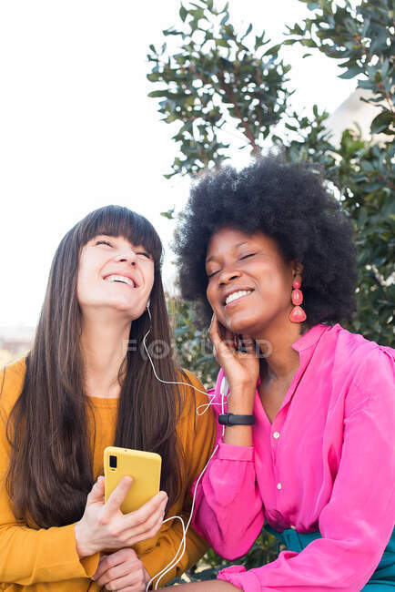 Cheerful multiracial LGBT couple of females listening to music in earphones and enjoying songs with closed eyes together in summer park — Stock Photo