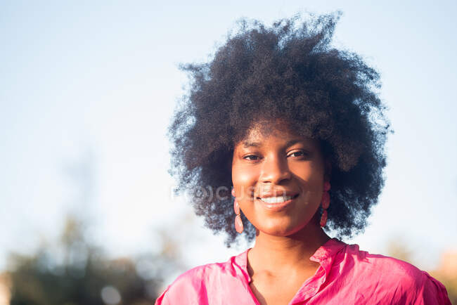 Charming African American female with curly hair smiling at camera while standing on sunny day in park — Stock Photo
