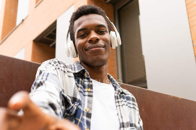 Dal basso di positivo hipster afroamericano maschile che ascolta musica in cuffia e si gode le canzoni mentre tende la mano verso la fotocamera in strada — Foto stock