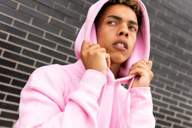Confident serious young curly haired hipster guy in pink hoodie looking away against brick wall — Stock Photo