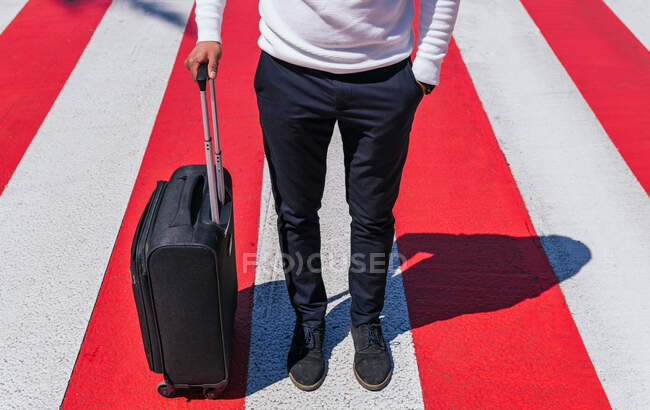 Сверху взрослый мужчина в элегантной повседневной одежде прогуливается с рукой в кармане и чемоданом через дорогу на современной солнечной улице — стоковое фото