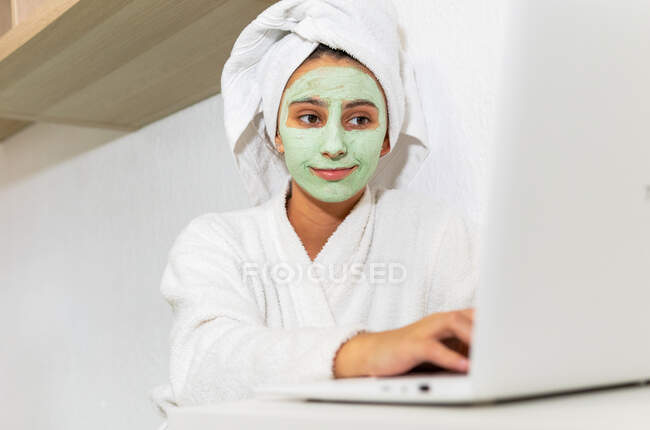Jeune femme avec masque de boue verte assis à la table et le netbook de navigation pendant la routine de soins de la peau le matin à la maison — Photo de stock