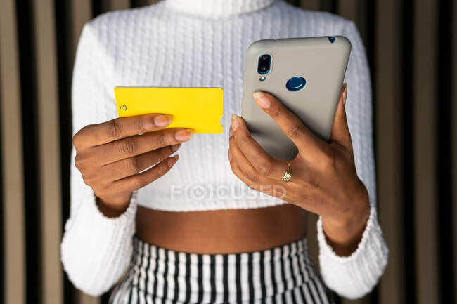 Нерозбірлива молода афроамериканська жінка в модному одязі, використовуючи кредитну картку і смартфон, щоб заплатити за онлайн-замовлення проти смугастих стін на вулиці — стокове фото