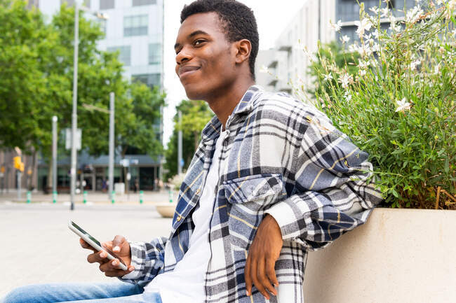 Felice maschio afroamericano elegante seduto sulla panchina navigando su smartphone in città e guardando altrove — Foto stock