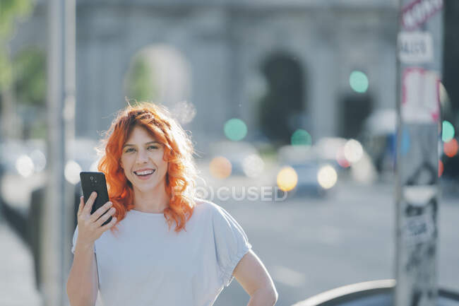 Alegre pelirroja mujer en la calle y mensajería en las redes sociales en el teléfono móvil - foto de stock