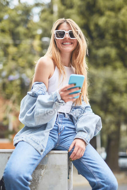 Trendy allegra donna in denim vestito seduto in strada e la navigazione sui social media sul telefono cellulare — Foto stock