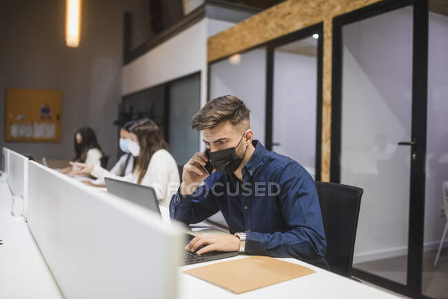 Empleado masculino en máscara escribiendo en el portátil y hablando en el teléfono inteligente mientras trabaja en el espacio de coworking con sus colegas - foto de stock