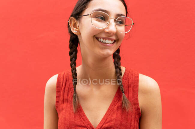 Fröhliche junge Frau mit Zopf-Frisur, während sie auf rotem Hintergrund in der Straße wegschaut — Stockfoto
