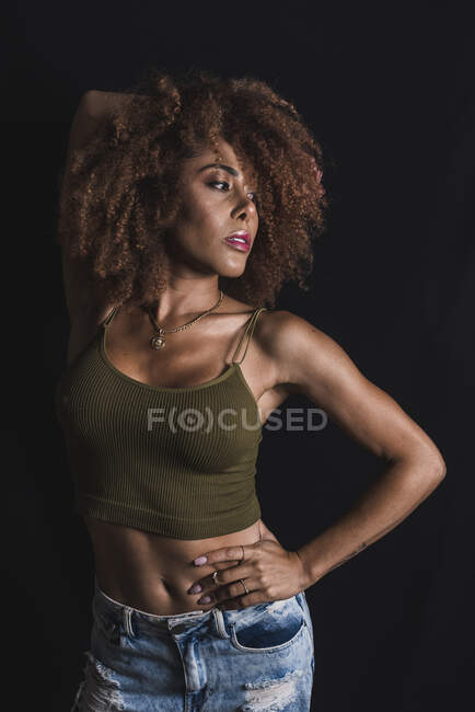 Mujer afroamericana de pelo rizado en top de moda y jeans de pie con la mano en la cintura sobre fondo negro en el estudio - foto de stock