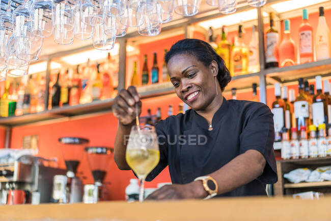 Sonriente mujer barman afroamericana mezclando cóctel frío colocado en el mostrador en el bar - foto de stock