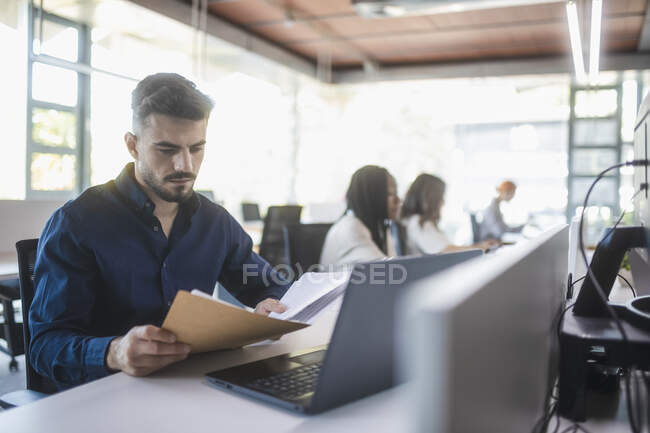 Empregado masculino concentrado sentado à mesa com laptop e lendo documentos enquanto trabalhava em um escritório espaçoso com colegas desfocados — Fotografia de Stock