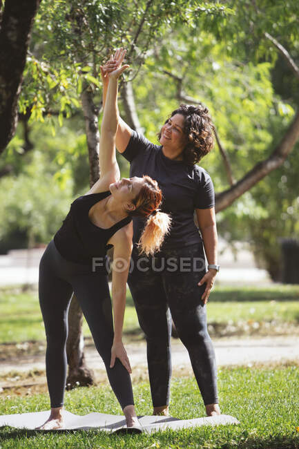 Ganzkörper junge barfüßige Anfängerin, die auf einer Matte auf dem Rasen steht und Trikonasana-Pose macht, während sie Yoga im grünen Park mit Hilfe eines ethnischen Personal Trainers praktiziert — Stockfoto