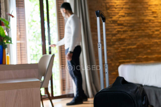Full body side view of adult man with luggage opening balcony door in hotel room — Stock Photo