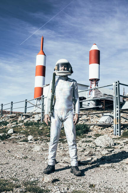 Homem de corpo inteiro em traje espacial em pé em solo rochoso contra cerca de metal e foguete listrado em forma de antenas no dia ensolarado — Fotografia de Stock