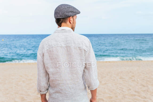 Vista trasera de un hombre irreconocible con ropa casual de moda y gorra caminando solo en la playa de arena hacia el mar ondeando mientras pasa las vacaciones de verano en la orilla del mar - foto de stock
