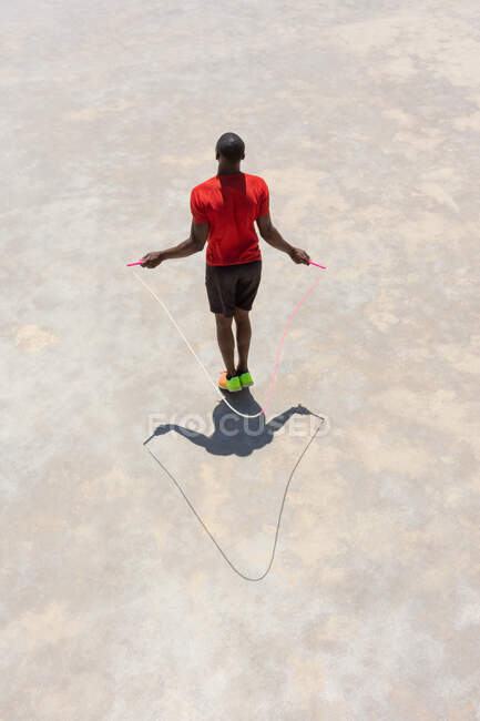 Сверху сзади вид безымянного афроамериканского спортсмена, прыгающего с каната во время тренировки в солнечный день на спортивной площадке — стоковое фото