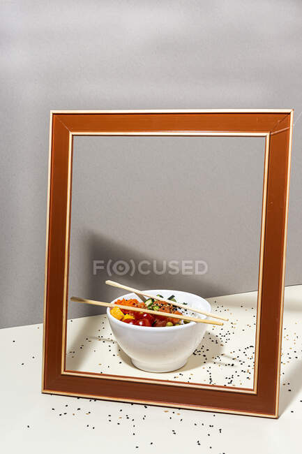 Високий кут білої миски зі смачною тарілкою і паличками, розміщеними за рамкою на столі, покритому кунжутом — стокове фото