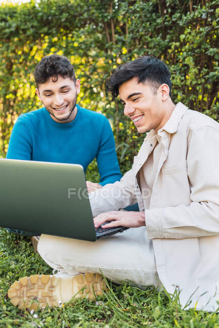 Colegas masculinos navegando netbook y trabajando en el proyecto de forma remota mientras se sienta en el césped en el parque - foto de stock