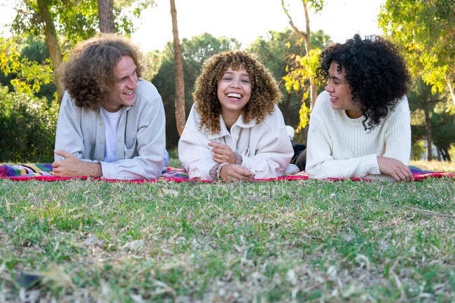 Diverse Freundinnen und Freunde mit lockigem Haar liegen auf einer bunten Decke auf der Wiese und unterhalten sich im Sommer — Stockfoto