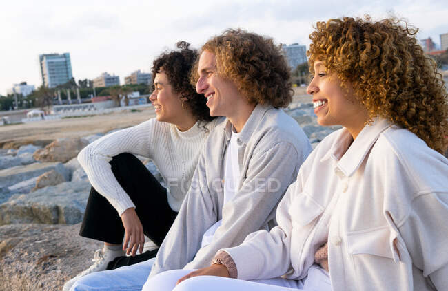 Side view of cheerful friends with curly hair sitting close against cityscape and rocky embankment in sunlight — Stock Photo