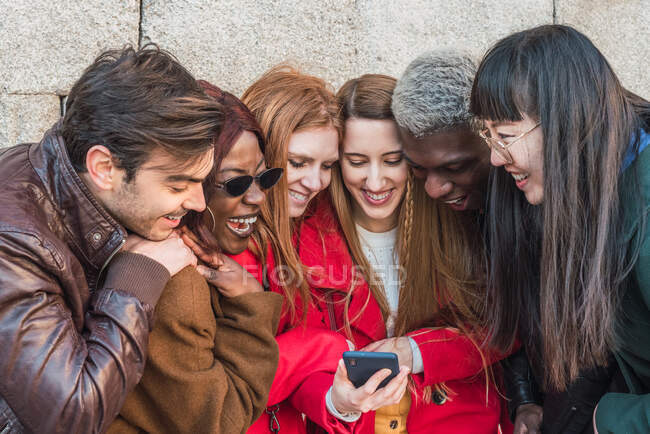 Empresa de amigos multiétnicos elegantes que se reúnem na rua e assistem vídeo engraçado no telefone celular juntos — Fotografia de Stock