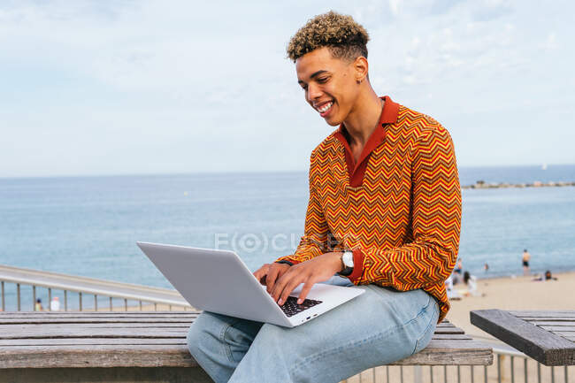 Positive young ethnic male student in stylish outfit typing on laptop keyboard while sitting on bench near sea on urban beach — Stock Photo
