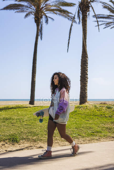 Полный вид со стороны тела женщины со скейтбордом в руке прогуливаясь по тротуару вдоль высоких ладоней против побережья и моря — стоковое фото