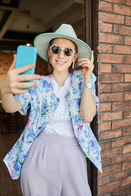 Cheerful young female in sunglasses and trendy apparel taking self portrait on cellphone against urban building — Stock Photo