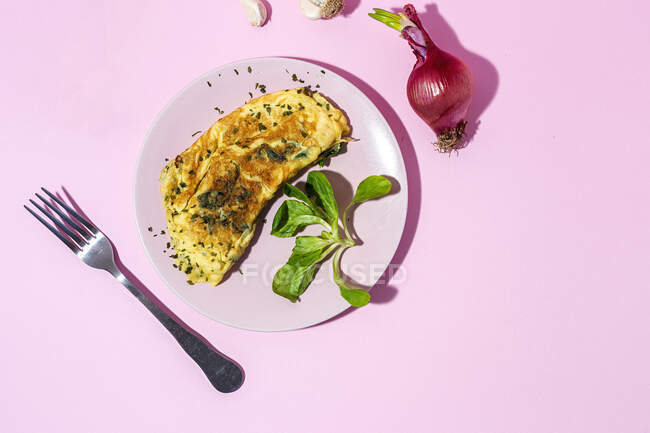 Sabrosa tortilla en plato contra ramitas de perejil fresco y cebolla roja con dientes de ajo sobre fondo rosa - foto de stock