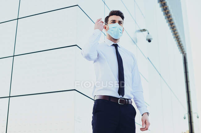 Снизу предприниматель-мужчина в формальной одежде с медицинской маской, смотрящий в город во время пандемии коронавируса — стоковое фото