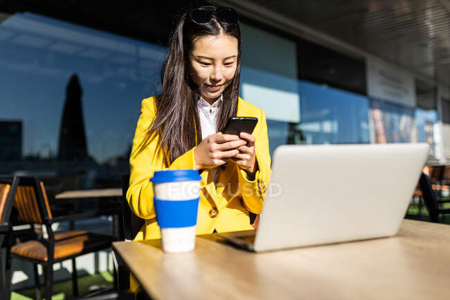 Азійська бізнес-жінка з жовтим пальто сидить за столом і п'є каву зі своїм розумним телефоном і ноутбуком. — стокове фото