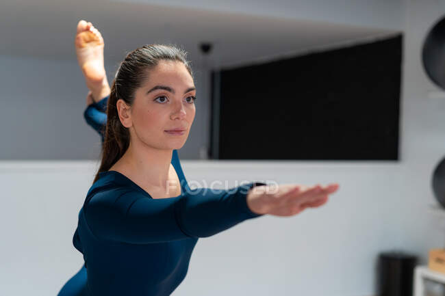 Vista lateral de una joven hembra delgada en ropa deportiva realizando la pose de Lord of the Dance mientras practica yoga en un gimnasio - foto de stock