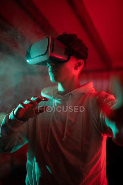 Человек в капюшоне во время использования VR очков и стоя в студии с красными неоновыми огнями — стоковое фото