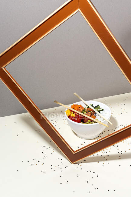 Alto ángulo de tazón blanco con sabroso plato poke y palillos colocados detrás del marco en la mesa cubierta con semillas de sésamo - foto de stock