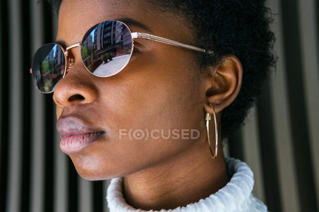 Vista lateral da jovem afro-americana em camisola elegante e óculos de sol olhando para longe, enquanto em pé na luz solar brilhante contra fundo preto — Fotografia de Stock