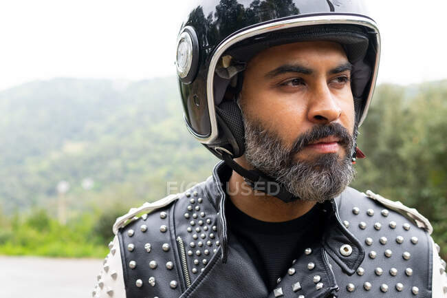 Middle aged bearded Hispanic male rider in trendy leather jacket with rivets and black helmet looking away while standing on road near lush green trees in nature — Stock Photo