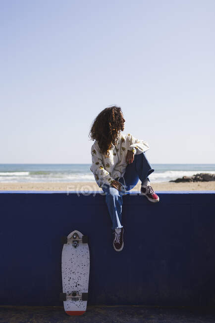 Полное тело положительной женщины сидит на перилах рядом со скейтбордом против песчаной набережной с морем в солнечную погоду — стоковое фото